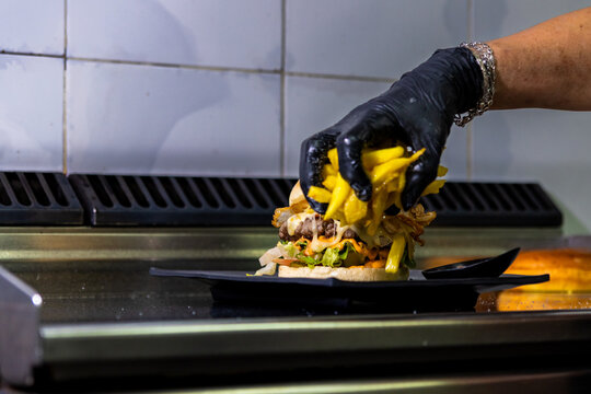 Hand in black glove adding french fries to a fresh burger on a dark plate