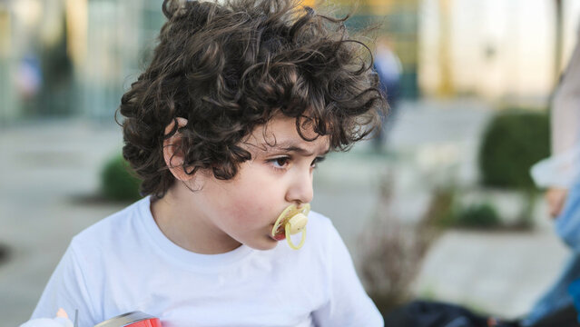 Young boy with curly hair using a pacifier looking away outdoors
