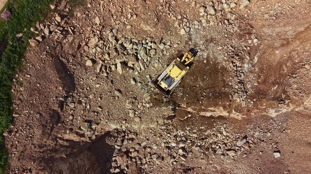 A bulldozer moves through a rocky landscape, pushing stones and dirt to prepare the ground for future development. The sun shines down on the work site, showcasing the machinery in action.