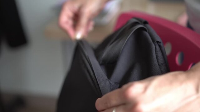 A close-up of a woman&rsquo;s hand unzipping a black rucksack. In the room, the woman unzips the rucksack, which is sitting on a pink chair.