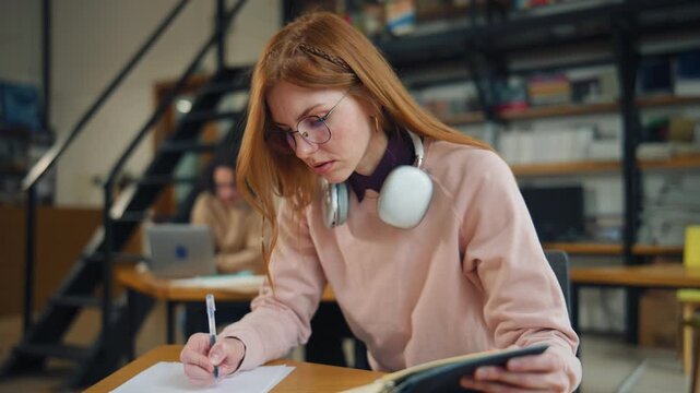 Woman writes notes in library while wearing headphones and seated at a desk during the day with another person in the background