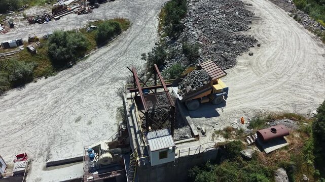 A large truck loads granite at a quarry site. Machinery and equipment are visible. Workers move around the area, which is filled with rock and debris. The sun is shining.