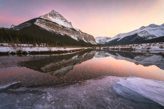View of the snow-capped peaks of the Canadian Rockies reflecting in the still water of a river with ice along the banks during a pink sunset Jasper, Alberta, Canada.
