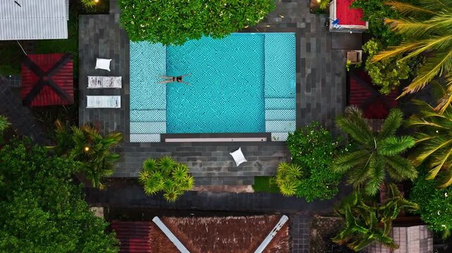 Top down aerial view of woman swimming in slow motion in turquoise tiled pool surrounded by lava stone deck and tropical garden villa at Gili Trawangan Lombok Indonesia