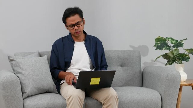 A young Asian businessman sits on the sofa in his living room while working on his laptop with an expression of disappointment, failure, stress due to bankruptcy.