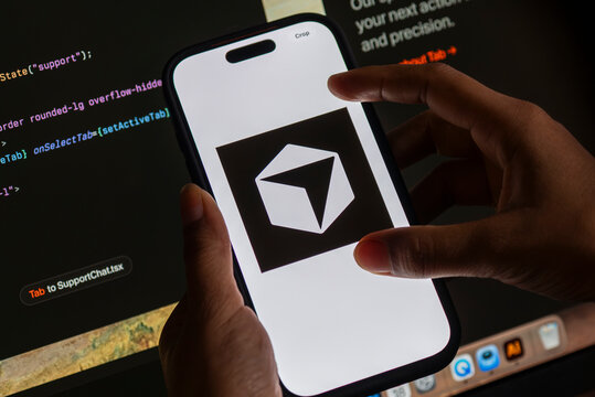 In this photo illustration, The Cursor AI logo is displayed on a smartphone screen in front of computer code and a software development interface