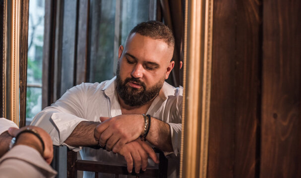 Confident plus size man with beard wearing a white unbuttoned shirt posing in a vintage interior, handsome larger male model looking at camera, body positivity and masculine style concept