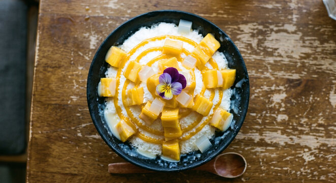 Korean mango bingsu shaved ice dessert with cubed yellow mango condensed milk and panna cotta in black slate bowl top down view