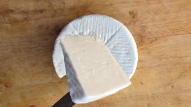 Top view of a person cutting a wheel of traditional white goat cheese on a rustic wooden board, gourmet food preparation and artisan cheese making concept in a Mediterranean kitchen
