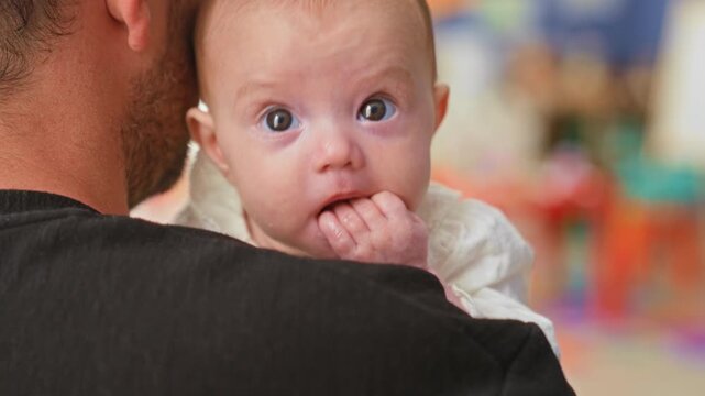 Baby girl held over adult shoulder with fingers in mouth and drool on chin in studio; quiet curiosity.