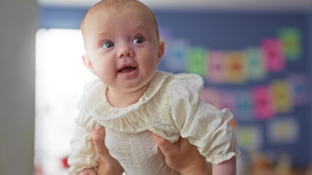 Baby girl face with chubby cheeks and wide eyes held by adult hands underarms in a colorful nursery building; gentle wonder.