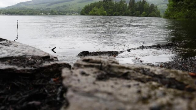 Rain on Loch Ard, Scotland, seen in a static close shot as droplets dimple the lake surface by a rocky shore with dark clouds and misty hills in the background.