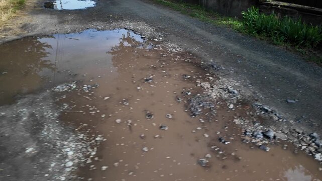 A cinematic 4K 24fps handheld shot of a muddy water puddle on a rough dirt road. Dynamic camera movement creates a realistic motion blur effect, capturing a gritty, realistic rural atmosphere.
