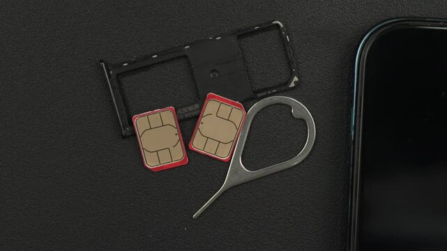 A SIM card tray, two red-edged SIM cards, and an ejector tool are placed next to a smartphone on a dark textured surface. Suitable for tech tutorials or mobile phone reviews.