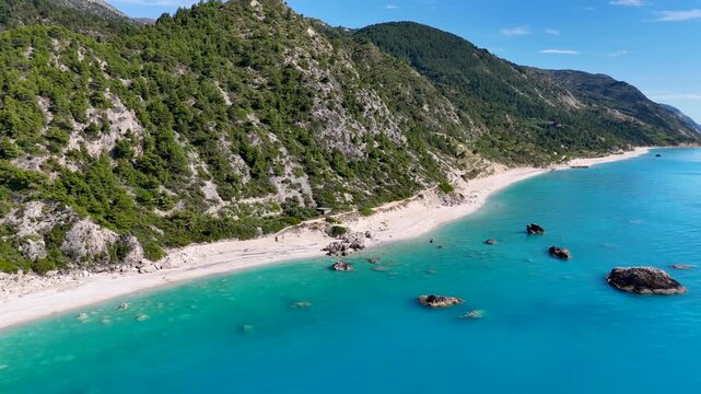Aerial drone footage flying along a secluded turquoise beach with crystal clear water, rocky shoreline and lush green mountains. Scenic coastal landscape on a sunny day.