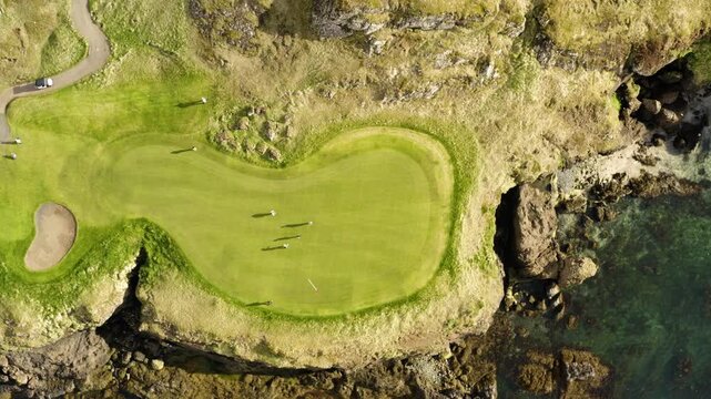 An amazing top-down aerial view of golfers playing on a beautiful green course situated on a rocky cliffside next to the clear ocean water.