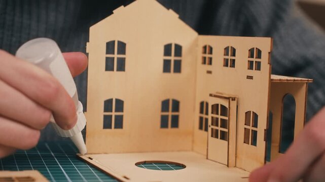 Attaching wall panel with glue during wooden house model construction. Hands assembling miniature home structure, DIY housing development, architectural crafting process, new residence concept