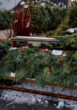 Evergreen wreaths with red berries displayed on a wooden stand in a snowy outdoor market.