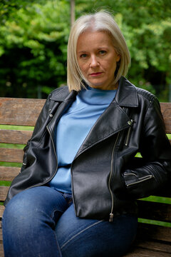 Woman in a leather jacket sitting on a wooden bench in a park setting.