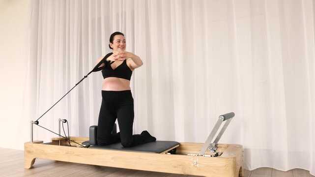 Pregnant woman using wooden pilates reformer with resistance straps exercise