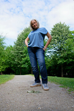 Woman in blue shirt and jeans standing on a park path surrounded by green trees.