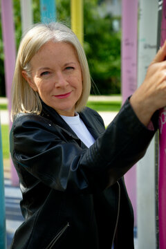 Woman in black jacket smiling while leaning on colorful poles in a sunny outdoor setting.
