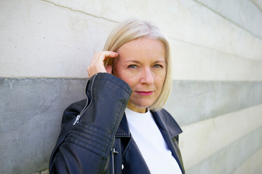 Mature woman in black leather jacket leaning against a modern concrete wall.