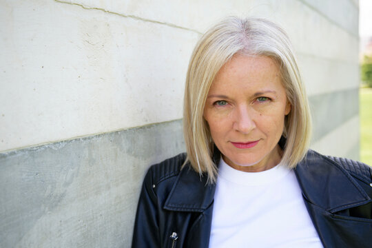 Blonde woman in a leather jacket poses confidently against a concrete wall.