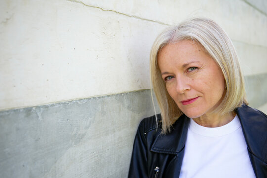 Woman with blonde hair and a leather jacket stands against a concrete wall, looking confidently.