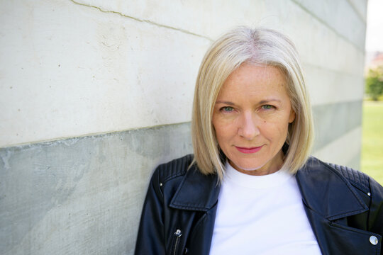 Blonde woman in a leather jacket leans against a white and gray concrete wall outside.