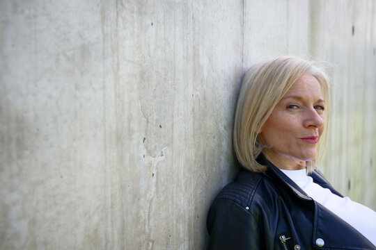 Woman with short blonde hair leaning against a textured concrete wall, wearing a leather jacket.