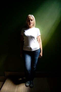 Woman in a white t-shirt and jeans stands against a green wall in dramatic lighting.