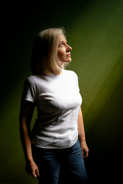 Mature woman in white t-shirt and jeans standing against a dark green background.