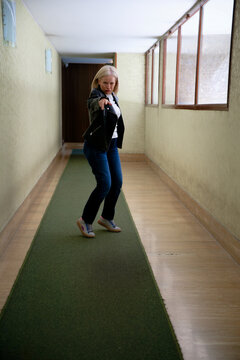 Woman in casual outfit playfully posing and pointing in a long hallway with green carpet.