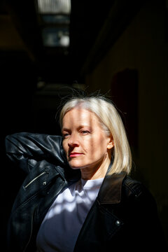 Woman with short blonde hair in leather jacket lit by sunlight indoors.
