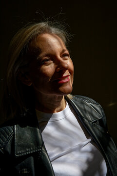 Mature woman in leather jacket and white shirt smiling subtly under dramatic lighting.