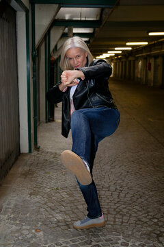 Woman in leather jacket and jeans striking a martial arts pose in an urban parking area.