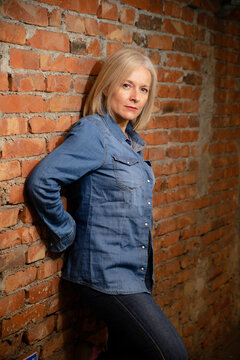 Mature woman in a denim outfit leans against a rustic brick wall, looking confidently at camera.