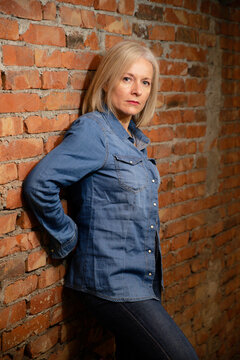 Mature woman in denim jacket poses against a rustic brick wall with a serious expression.
