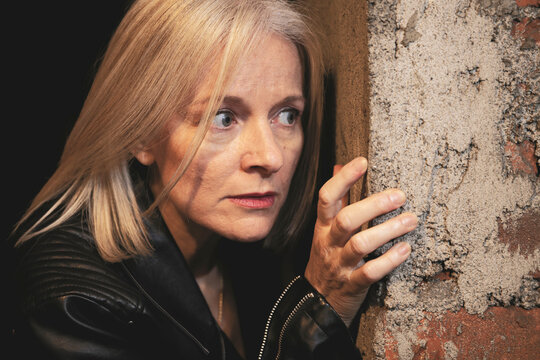 Woman with long blonde hair peering around a stone wall, wearing a black leather jacket.