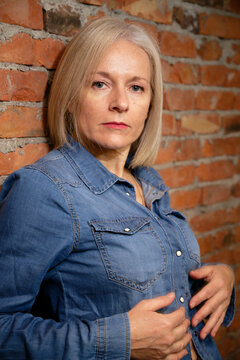 Woman in denim shirt leaning against a brick wall with a serious expression.
