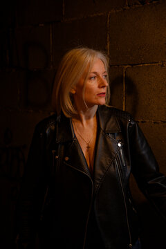 Woman in leather jacket stands thoughtfully against a shadowy brick wall with light on her face.