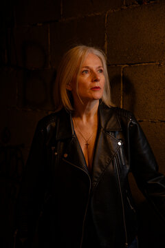 A woman with blonde hair in a black leather jacket stands against a dimly lit brick wall.