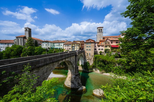 Stone bridge over a clear turquoise river with colorful historic buildings and a lush landscape.