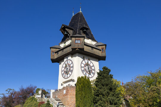 View of the historic Grazer Uhrturm clock tower with its large clock faces and wooden gallery under a clear blue sky in Graz, Styria, Austria.