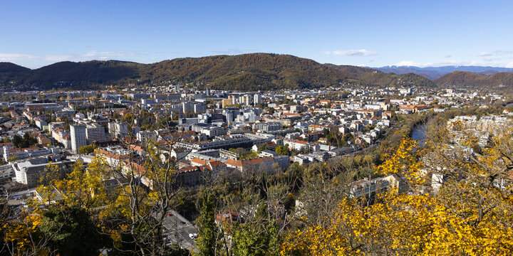 View of the city skyline with autumn foliage on trees, the Mur river, and rolling hills under a clear blue sky in Graz, Styria, Austria.