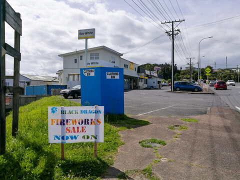 A street corner in Browns Bay features a fireworks sale sign, a blue clothing donation bin, parked cars, and local businesses under a partly cloudy suburban sky