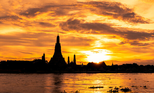Wat Arun Ratchawararam silhouetted against a dramatic orange sunset sky above the Chao Phraya River. The iconic Buddhist temple, cultural heritage and historic landmark of Bangkok, Thailand.