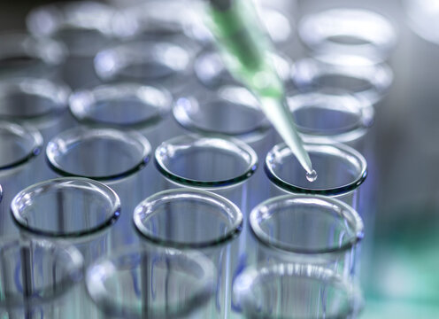 Droplet from pipette into rows of clear glass test tubes in a laboratory setting.