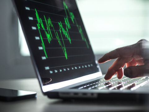 Hand typing on a laptop keyboard with stock market graph on screen